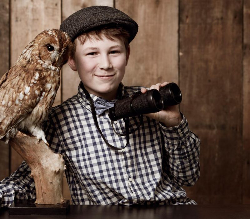 Ornithology is so much fun. Young boy in retro clothing wearing spectacles holding binoculars alongside an owl.