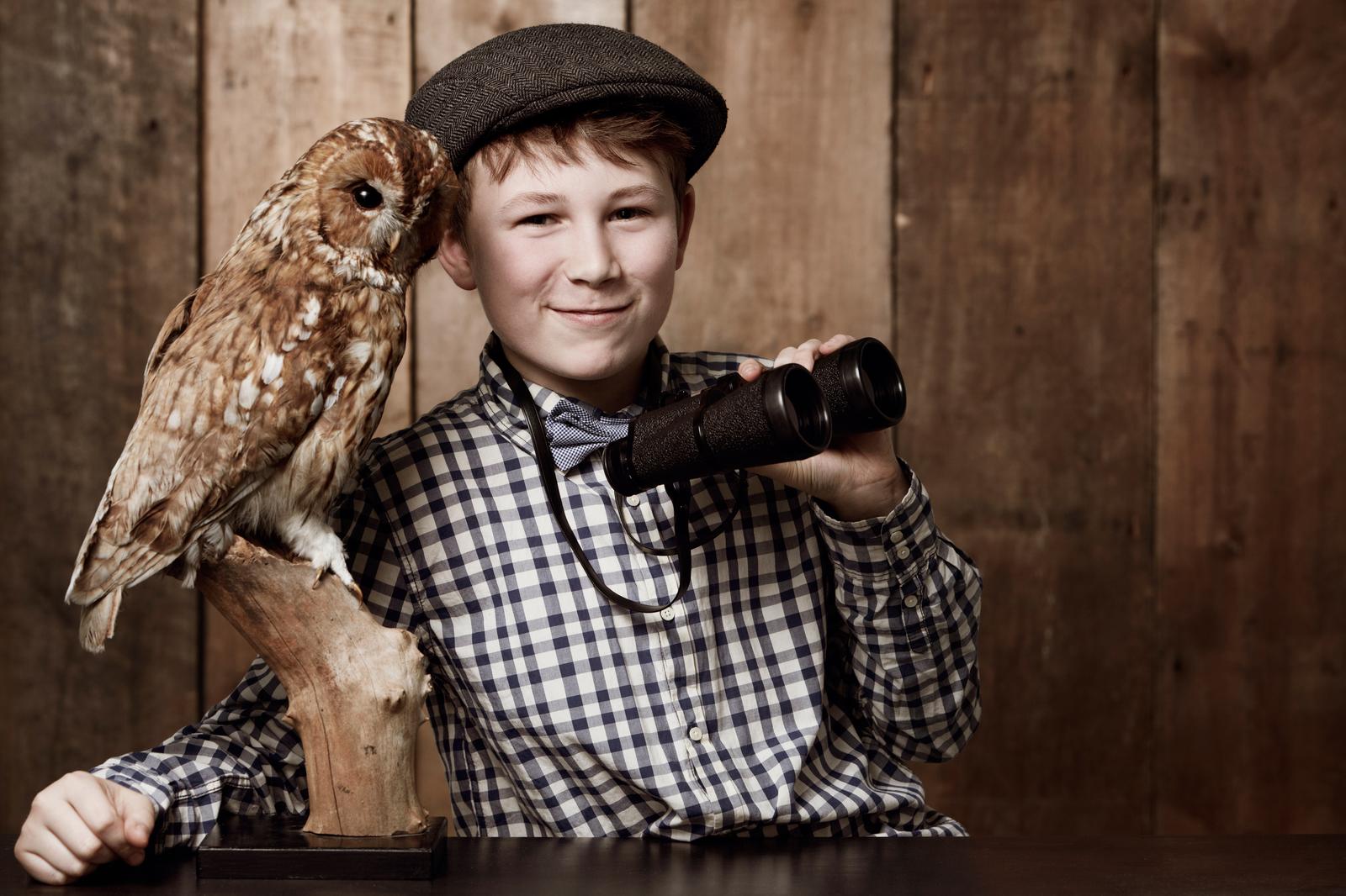 Ornithology is so much fun. Young boy in retro clothing wearing spectacles holding binoculars alongside an owl.
